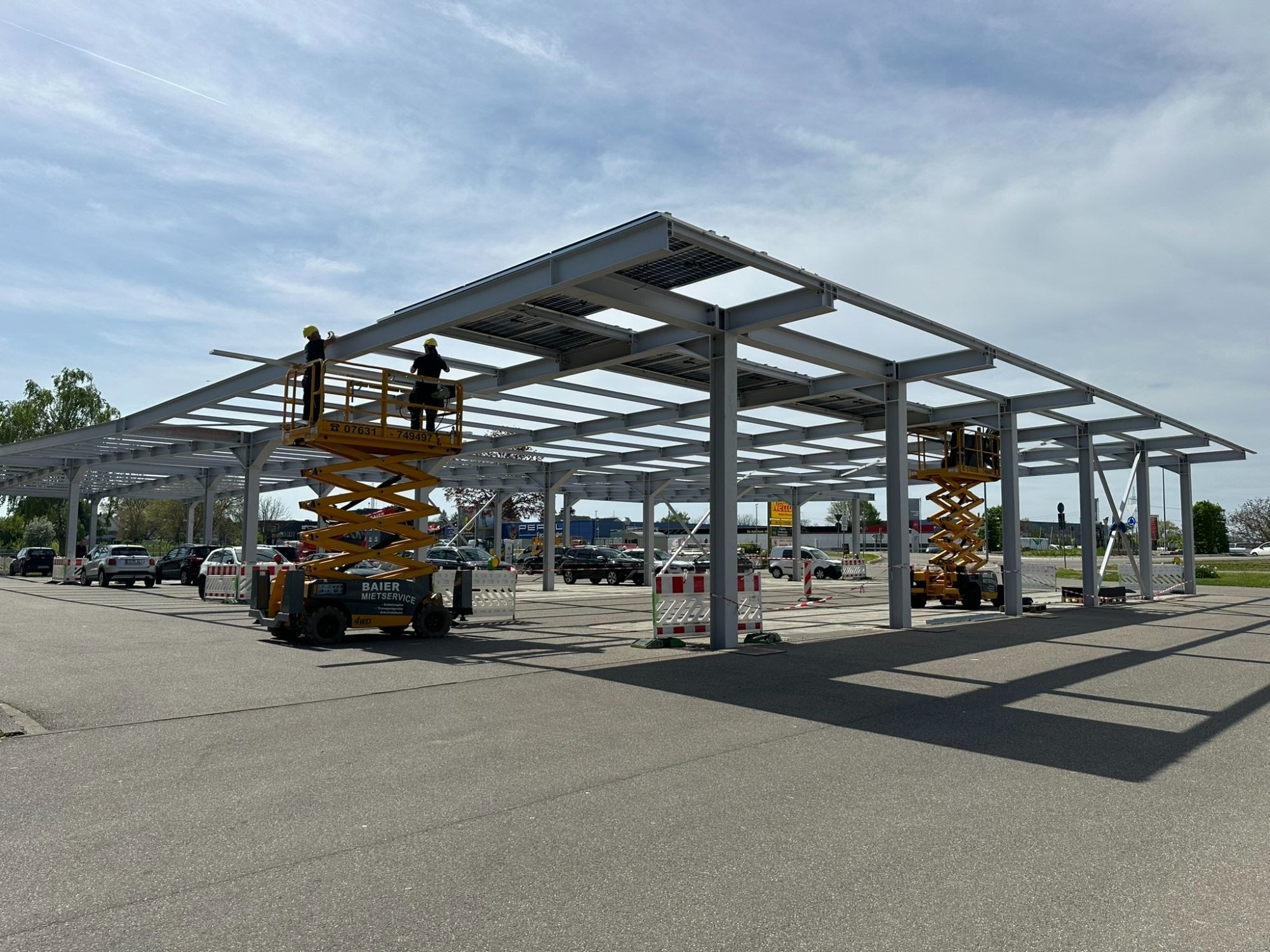 Bauphase Auggen: Stahlkonstruktion des PV-Carports im Aufbau, zwei Scherenbühnen montieren die Dachträger über dem Parkplatz