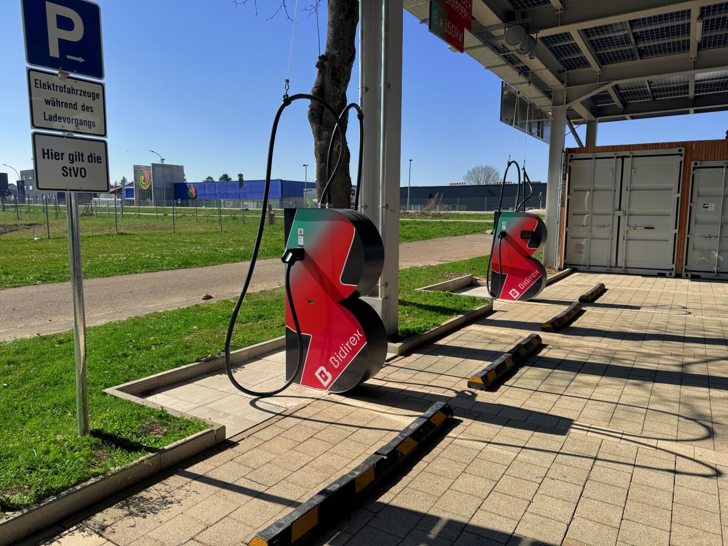 Zwei Bidirex-DC-Schnellladesäulen mit rot-grünem Markenwrap am Ladepark Auggen, dahinter Technik-Container und das Schild '8 × 150 kW Charger'