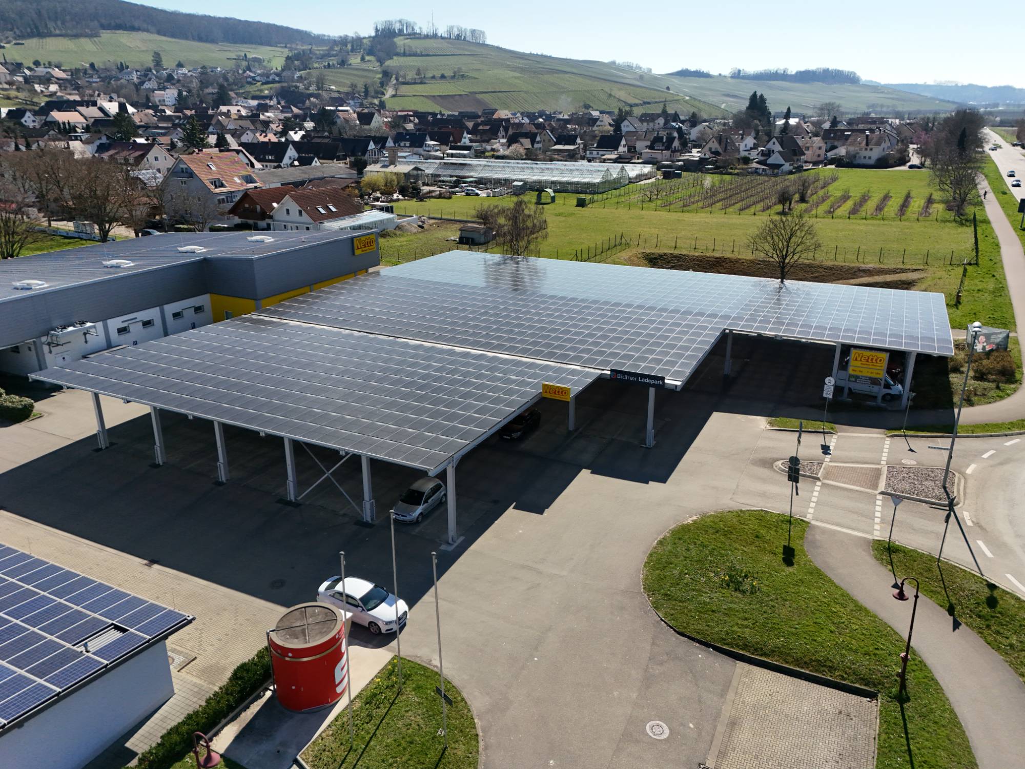 Drohnenaufnahme: Bidirex-Ladepark Auggen mit PV-Carports neben dem Netto-Markt, im Hintergrund das Dorf Auggen und das Markgräflerland